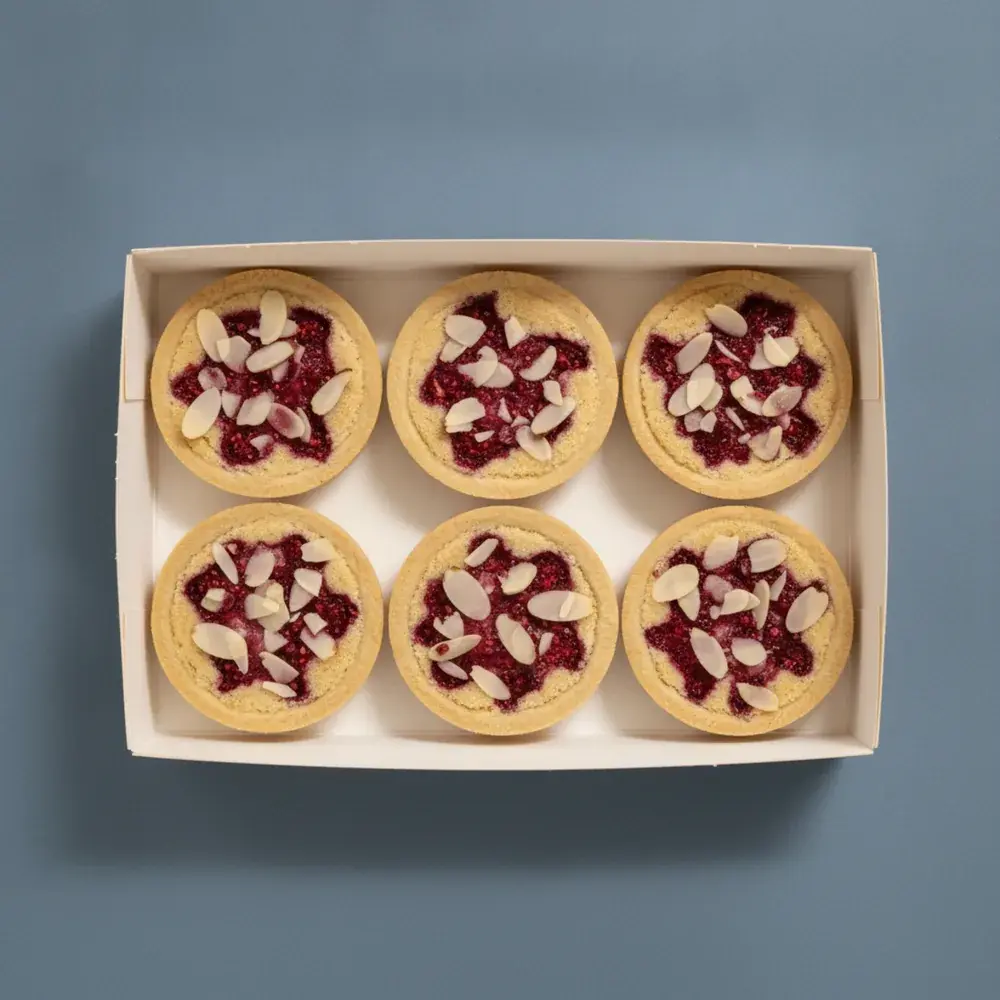 A box of six raspberry almond bakewell tarts, crushed fruit through baked frangipane, scattered with almond flakes