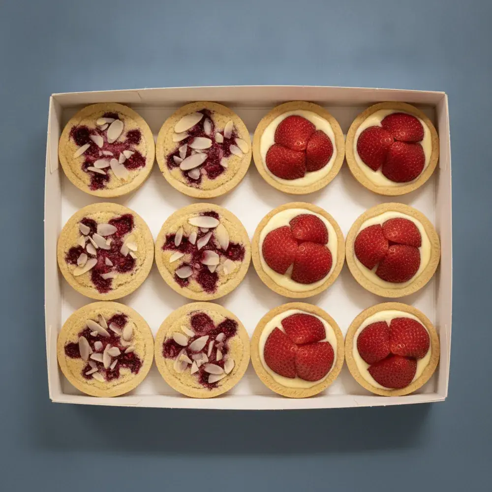An open box of twelve French raspberry almond bakewell and strawberry custard tarts, seen from above